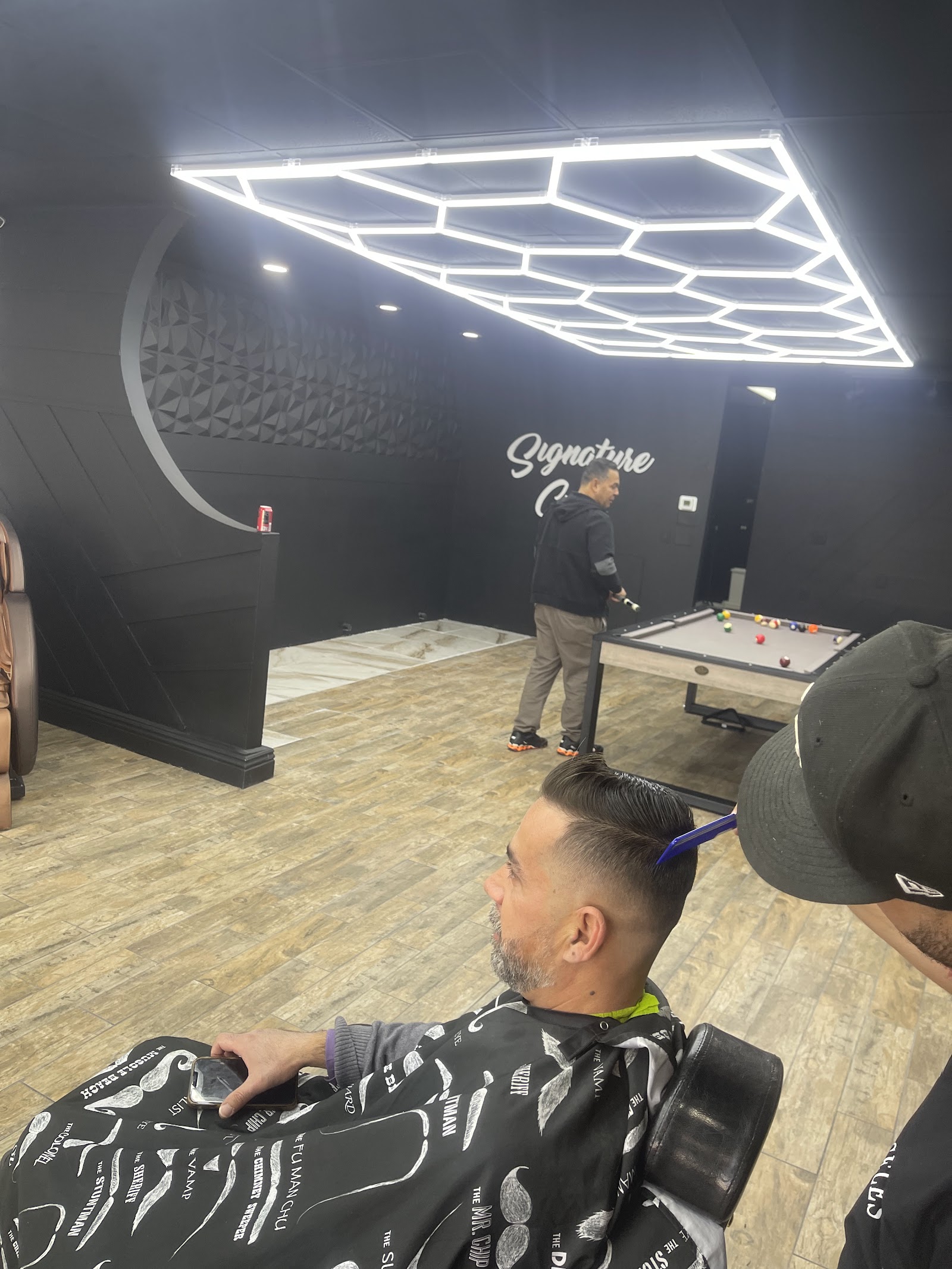 Signature Cuts Barbershop interior with hexagonal LED lighting, a pool table and the Signature Cuts wall sign while a client gets a haircut